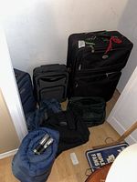Photo of luggage and sleeping bag lot against corner wall, showing three suitcases, two tote bags, rolled sleeping bag with flashlights inside