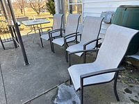 Rectangular patio table with glass top and 4 matching patio chairs lined up outside near a house wall on a patio surface