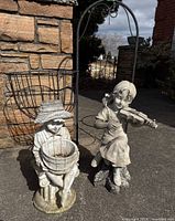 Shows two resin garden figurines and two wire planter stands outdoors by stone wall.