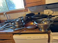 Photo of various pots, pans, and colanders arranged on a kitchen counter near sink and stove.