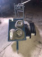 Photo showing blue metal wagon containing three utility tires on rims, black plastic vehicle ramps beside wagon, and metal-framed motorcycle lift at rear.