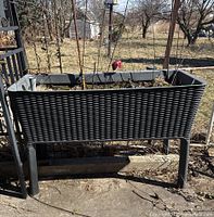 Front view of the dark grey resin planter with raised legs and rattan weave design, showing planter filled with soil and dry plant remains.