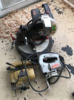 Photo showing Trade Master mitre saw along with two Black & Decker jigsaws on floor, cords visible and tangled.