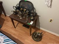 Full view of table with silver plate items including two tea/coffee pots, eight goblets, trays, creamers, sugar bowls, and utensils arranged on a wooden table and floor.