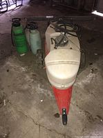 Overview of three handheld sprayers and one large wheeled sprayer cart with white tank and red base on concrete floor in garage setting.
