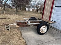 Side view of heavily used metal trailer frame with two wheels and wooden plank supports outside a barn.