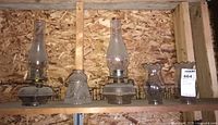 Shelf holding five vintage oil lamps and glass lamp shades of varying height and form in a garage area