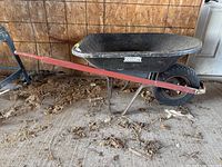 Side view of the Jobmate wheelbarrow showing black plastic tub, red handles, and single pneumatic tire on metal frame in a garage with wood wall and concrete floor.