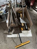Wooden wagon filled with various yard tools including shovels, pitchforks, a hoe, a rake, and a small yellow push broom. Tools show signs of rust and wear, indicating frequent outdoor use.