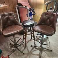 Two brown tufted faux leather bar chairs with metal footrests and bases, and a black wooden round stool between them.