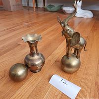 Photo showing three brass pieces: elephant figurine on a sphere, engraved vase with fluted rim, and a plain brass sphere on a wooden floor.