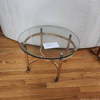 Side view of brass and glass side table on wooden floor, showing the three-legged brass frame with casters and thick clear glass round top.