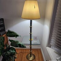 Full view of floor lamp with beige pleated shade, chrome and brass column with glass table, located in corner next to window and plants