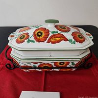 Stacked view of the two ceramic casseroles with poppy flower design on black wrought iron stand on red cloth.