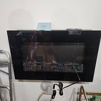 Electric fireplace mounted on wall with black frame and glass front. Visible decorative burning logs inside. Remote control with power cord placed below.