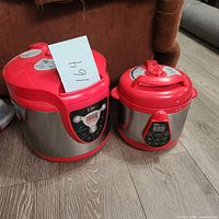 Two Elite electric pressure cookers of different sizes side by side on floor showing stainless steel bodies and red lids with digital controls