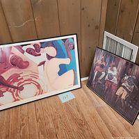 Two framed art pieces leaning against a wooden wall, one by Bernard Hoyes and one by Buchi Upjohn.