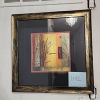 Photo shows the framed art piece by Don Li-Leger hung on a wall with a beige background. Frame is mottled gold/black, square shape, with mat surrounding the print inside.