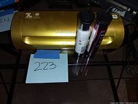 Gold Glaminator embossing and transfer machine with two rolls of transfer foil placed beside it on glass table, with handwritten lot number '223' on blue paper in front.