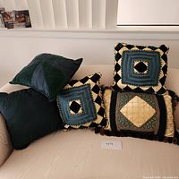 5 pillows displayed on a beige couch: 2 solid dark green pillows, 2 patterned square pillows with green, creme, blue and black colors, and 1 larger rectangular tapestry style pillow with fringes.