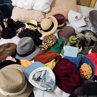 View of various hats and caps spread on a bed including fedoras, bucket hats, berets, baseball caps, and others.