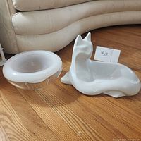 Both the large ceramic white cat figurine and the white ceramic bowl displayed on wooden floor next to a sofa.