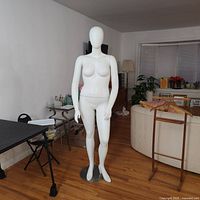 Full front view of a white female mannequin standing on a round metal base in a room with hardwood floors.