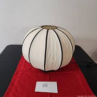 Front view of cocoon-shaped cream/beige fabric lampshade with black piping on a dark wood base placed on a red cloth.