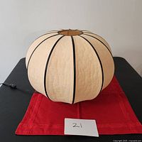 Cocoon-shaped pinkish cream fabric lamp shade with black piping, round design resting on a red cloth with a piece of paper marked '21' to indicate lot number or identification.