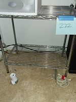 Metal wire shelving unit with three shelves, chrome finish, photographed from front showing full shelving unit with some items on floor nearby unassociated to shelving.