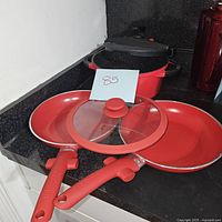 Photo of two red frying pans stacked with a glass lid on top, showing red handles and non-stick surfaces.