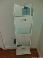 Front view of white wall organizer with three pull-out drawers, each with a metal label holder, positioned against a wall near a floor vent.