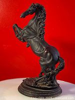 Side and front view of black resin horse statue rearing on hind legs on wooden base against red background