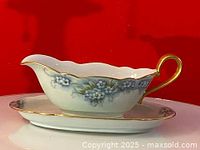 Gravy boat and underplate displayed on reflective surface against red background, showing front and top angles highlighting floral design, gold rims, and handle detail.