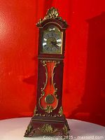 Front view of the miniature Comtoise style Schmidw grandfather clock showing the wooden case with ornamental golden trim and the clock face with roman numerals and decorative center.