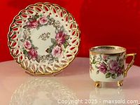 Photo of porcelain floral teacup with pink and purple flowers, gold trim, and footed base beside matching saucer with lattice pierced edge and floral design on a reflective surface