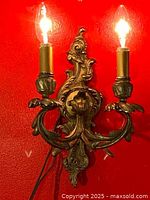 Full front view of the French Louis XV gilt bronze ormolu wall sconce mounted on a red wall with both lights illuminated.