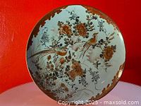 Full view of round Kutani pottery plate against red background showing intricate hand painted floral and bird design with brown border