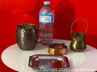 Four metal items on a white round table against a red background. Items shown are a hammered brass mug, small copper dish, small brass bucket, and a hammered silver plate tray.