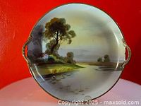 Top view of the round handled cake plate with hand-painted countryside with house, trees, and water scene with gold trim on handle edges and rim.