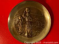 Full view of the round solid brass plate showing the embossed design of two Native American women in traditional dress carrying pottery on their heads, set against a textured landscape background.