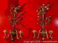 Front view of a pair of gold-tone metal wall sconces with detailed leaf motifs and dual candle holders attached to curved arms.
