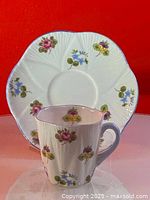 Teacup and matching saucer displayed on pink background showing floral pattern and scalloped edges with blue rim.