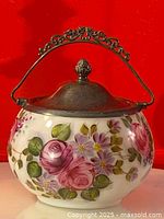 Front view of biscuit barrel showing detailed pink and purple flower painting, silver plated lid and decorative handle against red background.