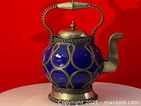 Front view of vintage blue cobalt ceramic teapot with silver handle, spout and detailed metal overlay on body, lid, and base against red background.