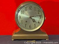 Front view of the clock on a stepped base with silver dial and Arabic numerals, key positioned in front.