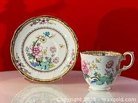Photo showing full teacup and saucer pair with floral design and gold trim on red background.