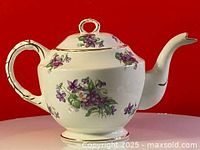 Front side view of the white porcelain teapot with violet floral pattern against a red background.