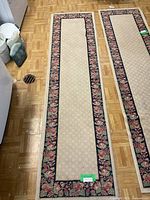 Two long beige carpet runners with black borders and floral patterns laid out on a parquet floor near laundry items.