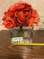Front view showing large artificial orange flowers in a clear glass square vase. A yellow measuring tape indicates vase dimensions around 6 inches tall.